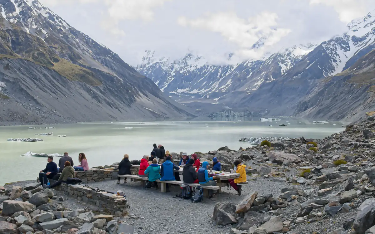 A group of visitors sitting at picnic tables beside a glacial lake below the snow-covered peaks of Aoraki Mount Cook, New Zealand