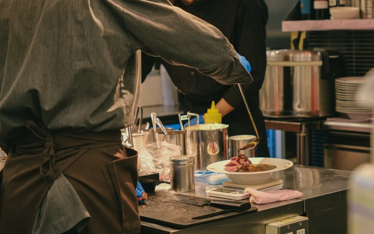 Chef ladling sauce over a plated dish during service in a restaurant kitchen
